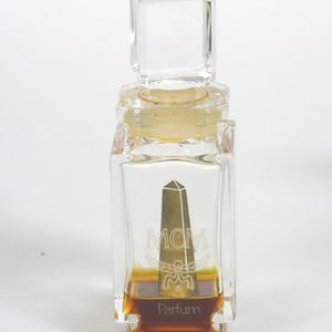 Pode incluir: Um frasco de perfume de vidro transparente com uma tampa retangular. O frasco contém um líquido castanho e apresenta um design de obelisco dourado com as letras "MCM" e a palavra "Parfum".