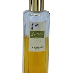 May include: A rectangular perfume bottle with a gold cap and a label that reads "Sortilège PARFUM DE TOILETTE LE GALION." The bottle is clear with a ribbed design and contains a golden-coloured liquid.