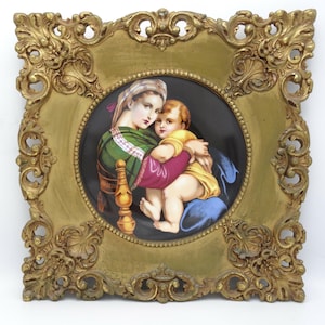May include: A square, ornate gold-colored frame surrounds a circular porcelain plaque featuring a painted scene of a person in a green and purple garment holding a child in a yellow shirt against a solid black background with a beaded border.