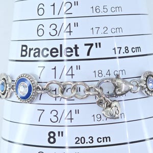 May include: Silver bracelet with blue and clear gemstone accents. The bracelet is approximately 7 inches (17.8 cm) long and features a heart-shaped charm. The bracelet is displayed on a white measuring guide.