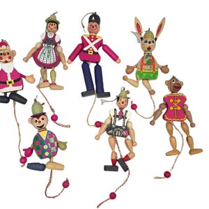 Può includere: Una collezione di sette ornamenti in legno a molla che raffigurano vari personaggi, tra cui un Babbo Natale, un soldato, un coniglio e figure in abiti tradizionali. Ogni figura in legno dipinto ha arti mobili azionati da una cordicella.
