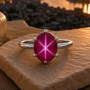 May include: A silver ring featuring a large, oval-cut ruby gemstone. The ruby displays a six-rayed star effect. The ring is set on a textured, light brown surface, with a blurred cityscape and sunset in the background.
