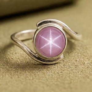 May include: A sterling silver ring featuring a round, pink star sapphire gemstone. The gemstone displays a six-rayed star pattern. The ring has a swirling design on the band, adding a touch of elegance to the jewelry.