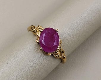 Könnte beinhalten: Ein goldener Ring mit einem ovalen Rubin. Der Ring hat ein detailliertes, verziertes Banddesign, möglicherweise floral oder blattartig. Der Rubin hat eine tiefe, satte rote Farbe. Der Ring ist vor einem neutralen Hintergrund platziert.