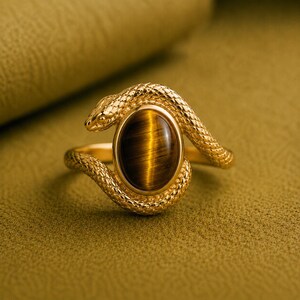 Può includere: Un anello color oro caratterizzato da un design a serpente testurizzato che avvolge una gemma centrale a forma ovale in occhio di tigre. La fascia in metallo presenta un motivo a scaglie dettagliato, mentre la pietra marrone mostra una banda di luce chatoyante.