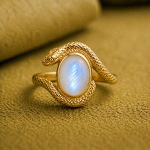 May include: A gold ring featuring a textured snake motif that wraps around a central oval-shaped moonstone. The snake has detailed scales and a defined head, while the moonstone displays a soft, iridescent blue glow within a smooth gold bezel.