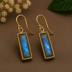 Puede incluir: Un par de pendientes colgantes de tono dorado con piedras de labradorita rectangulares de superficie facetada. Las piedras muestran una iridiscencia azul vibrante engastadas en un marco de metal liso, suspendidas de ganchos dorados.