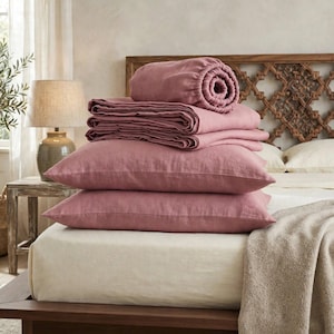 May include: A stack of dusty rose colored bedding items including a fitted sheet, a flat sheet, and two rectangular pillowcases. The bedding is made of a textured fabric and is arranged on top of a cream colored mattress on a wooden bed frame.