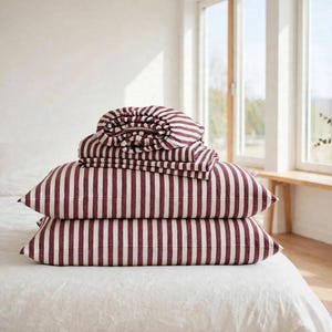 May include: A stack of bedding featuring two rectangular pillows and a folded flat sheet, all decorated with a vertical striped pattern in deep burgundy and off-white. The fabric has a visible woven texture throughout the entire set.