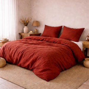 Puede incluir: Una habitación con una cama vestida con una funda nórdica y almohadas de color óxido. La cama está sobre una alfombra beige, y la habitación tiene una paleta de colores neutros con vigas de madera en el techo. Hay un jarrón con flores secas y una lámpara pequeña.