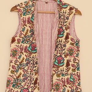 May include: A sleeveless vest with a floral pattern in teal, pink, and yellow on an off-white background. The interior lining features vertical red and white stripes. The vest is hanging on a wooden hanger.
