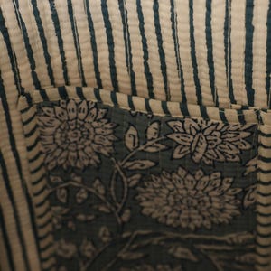 May include: Close-up of a fabric with a striped pattern in off-white and teal. A square patch with floral designs in teal and white is sewn onto the fabric, creating a pocket. The fabric appears to be quilted.