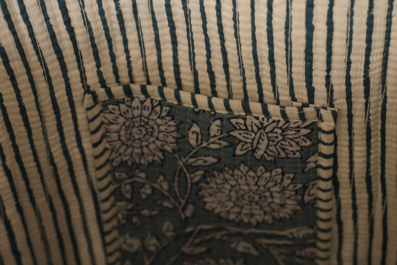 May include: Close-up of a fabric with a striped pattern in off-white and teal. A square patch with floral designs in teal and white is sewn onto the fabric, creating a pocket. The fabric appears to be quilted.