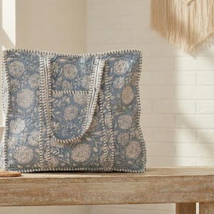 May include: A large tote bag with a blue floral pattern. The bag has a rectangular shape with a wide opening and two handles. The floral design is in white and the bag is sitting on a wooden bench.