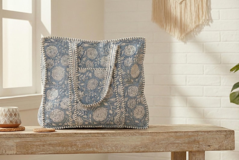 May include: A large tote bag with a blue floral pattern. The bag has a rectangular shape with a wide opening and two handles. The floral design is in white and the bag is sitting on a wooden bench.