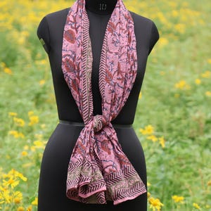 May include: A pink scarf with a floral pattern and geometric border is draped on a black mannequin. The scarf features a repeating design of red flowers and green leaves. The background is a field of yellow flowers.