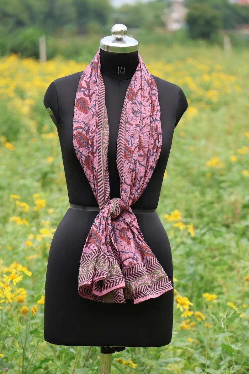 May include: A pink scarf with a floral pattern and geometric border is draped on a black mannequin. The scarf features a repeating design of red flowers and green leaves. The background is a field of yellow flowers.