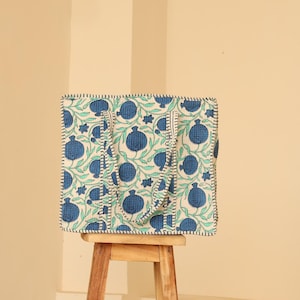 May include: A square tote bag with a white background and a blue pomegranate pattern. The bag has two handles and is sitting on a small wooden stool. The bag's edges are outlined with black stitching.