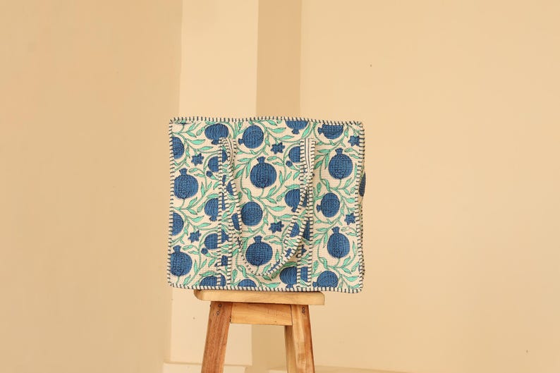 May include: A square tote bag with a white background and a blue pomegranate pattern. The bag has two handles and is sitting on a small wooden stool. The bag's edges are outlined with black stitching.