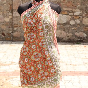 May include: An orange and green floral print sarong is draped over a mannequin. The sarong features a repeating pattern of flowers and leaves in green and white on an orange background. The edges have a striped border.