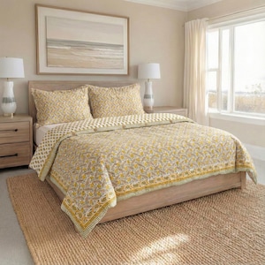 Puede incluir: Una cama con un edredón con estampado floral amarillo y fundas de almohada a juego. La ropa de cama está sobre una estructura de cama de madera clara, con una alfombra de fibra natural debajo. Dos lámparas se encuentran en las mesitas de noche y una pintura de paisaje enmarcada cuelga sobre el cabecero.