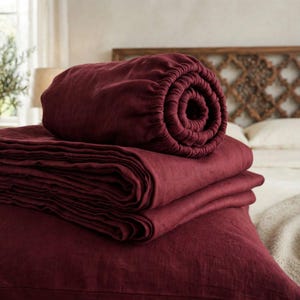 May include: A deep burgundy fitted sheet rolled into a cylinder, resting atop a stack of folded matching burgundy bed sheets. The fabric has a visible, textured weave and a matte finish, showcasing a rich, dark red color throughout the set.