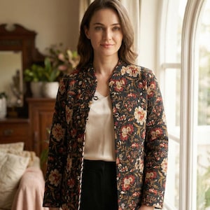 May include: A black quilted jacket with a floral pattern in red, orange, and cream. The jacket has a mandarin collar and is worn over a white top and black pants. The background includes a window and interior decor.