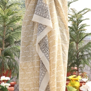 Pode incluir: Um tecido creme com um padr&atilde;o amarelo repetido e uma borda contrastante em preto e branco. A embalagem &eacute; drapeada sobre um manequim, mostrando seu design e textura. O tecido parece leve e adequado para climas quentes.