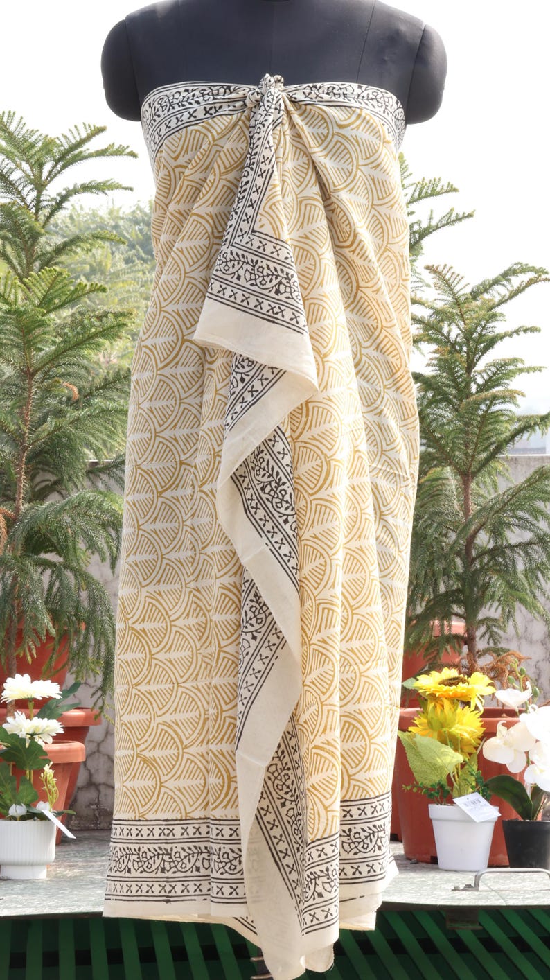 Pode incluir: Um tecido creme com um padr&atilde;o amarelo repetido e uma borda contrastante em preto e branco. A embalagem &eacute; drapeada sobre um manequim, mostrando seu design e textura. O tecido parece leve e adequado para climas quentes.