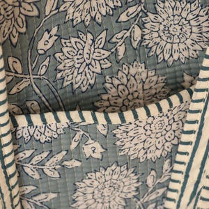 May include: Close-up of a quilted fabric with a floral pattern in white and teal. The fabric features large stylized flowers and leaves. Vertical stripes in teal and white border the edges, and a pocket is visible.