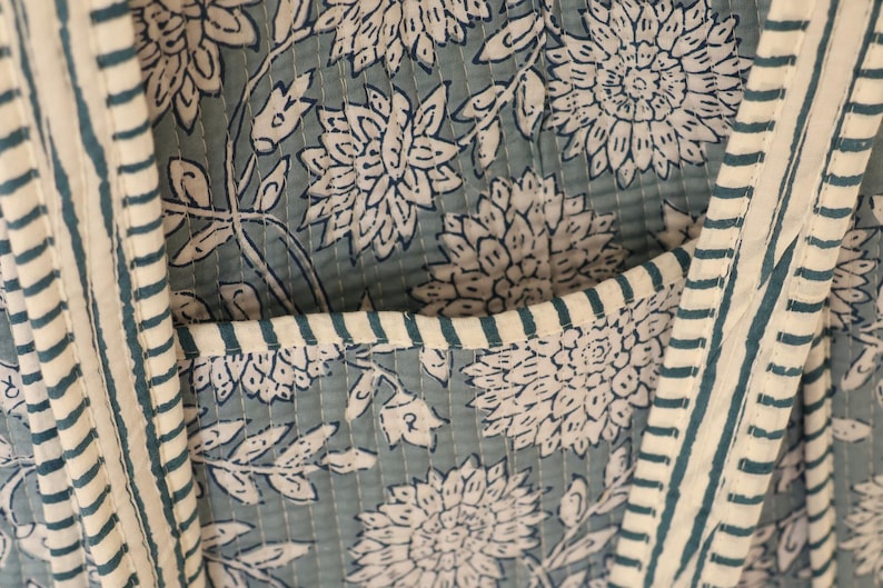 May include: Close-up of a quilted fabric with a floral pattern in white and teal. The fabric features large stylized flowers and leaves. Vertical stripes in teal and white border the edges, and a pocket is visible.
