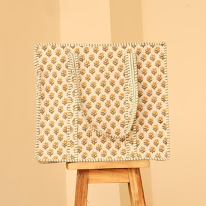 May include: A square tote bag with a floral pattern of yellow flowers on a cream background. The bag has two green straps and is sitting on a small wooden stool. The edges of the bag are outlined with a green stitch.