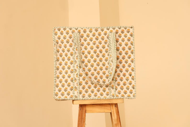 May include: A square tote bag with a floral pattern of yellow flowers on a cream background. The bag has two green straps and is sitting on a small wooden stool. The edges of the bag are outlined with a green stitch.
