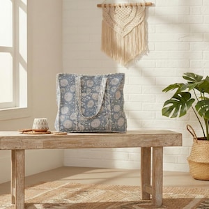 May include: A blue tote bag with a floral pattern sits on a wooden bench. The bag has white trim and handles. A small ceramic cup and a coaster are also on the bench. A potted plant and a macrame wall hanging are in the background.