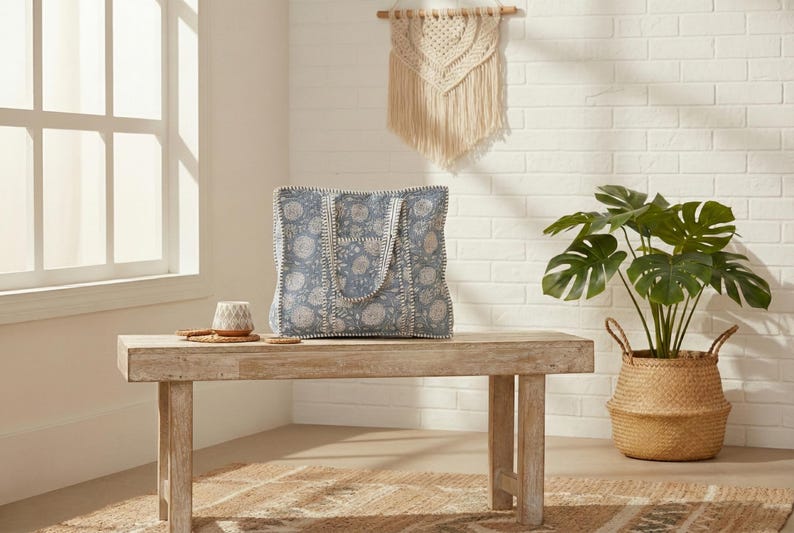 May include: A blue tote bag with a floral pattern sits on a wooden bench. The bag has white trim and handles. A small ceramic cup and a coaster are also on the bench. A potted plant and a macrame wall hanging are in the background.