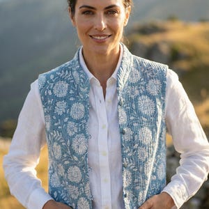 May include: A light blue quilted vest with a floral design, worn over a white shirt and dark jeans. The vest features a button closure and pockets. The image is set against a blurred outdoor backdrop.