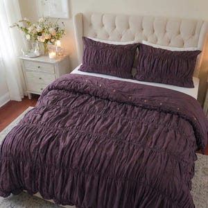 Könnte beinhalten: Eine pflaumenfarbene Bettdecke und zwei passende Kissenbezüge mit gerafftem Stoffdesign. Das Bettwäsche-Set befindet sich auf einem Bett mit einem cremefarbenen, knopfgetufteten Kopfteil. Der Raum hat eine neutrale Farbpalette mit einem Nachttisch und Blumen.
