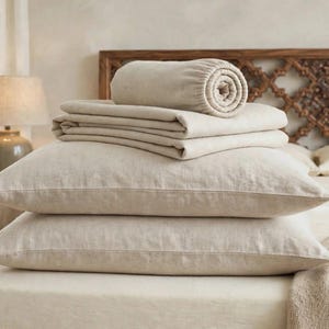 Puede incluir: Un juego de ropa de cama beige que incluye dos almohadas rectangulares apiladas, una sábana encimera doblada y una sábana bajera enrollada. La tela tiene una textura tejida visible en un tono beige neutro para la cama.