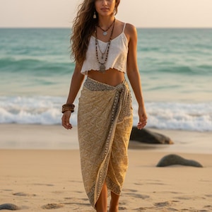 Pode incluir: Uma mulher com um top branco e um sarongue bege com uma borda estampada em preto caminha em uma praia arenosa. Ela usa colares e pulseiras em camadas. O oceano e o c&eacute;u s&atilde;o vis&iacute;veis ao fundo.