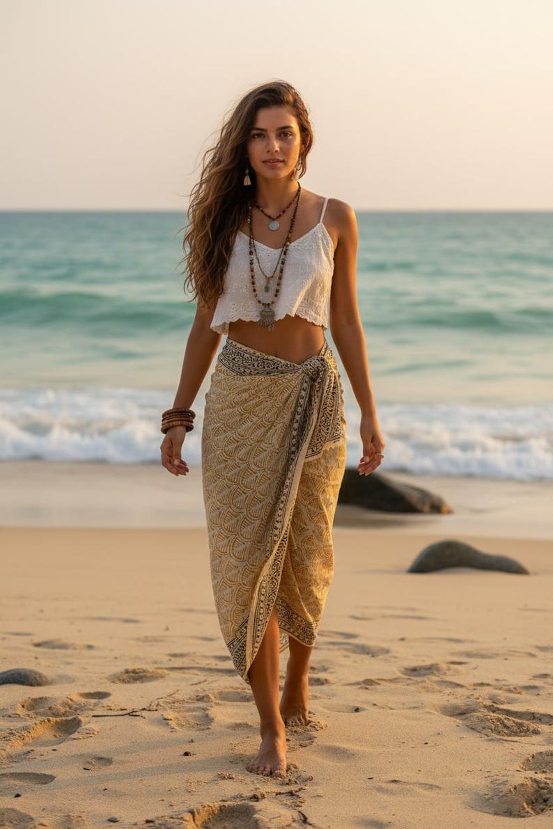 Pode incluir: Uma mulher com um top branco e um sarongue bege com uma borda estampada em preto caminha em uma praia arenosa. Ela usa colares e pulseiras em camadas. O oceano e o c&eacute;u s&atilde;o vis&iacute;veis ao fundo.