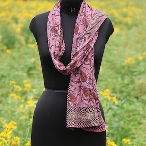 May include: A pink scarf with a floral pattern and a geometric border is draped on a black mannequin. The scarf features a repeating design of leaves and flowers in shades of red, blue, and brown. The background is a field of yellow flowers.