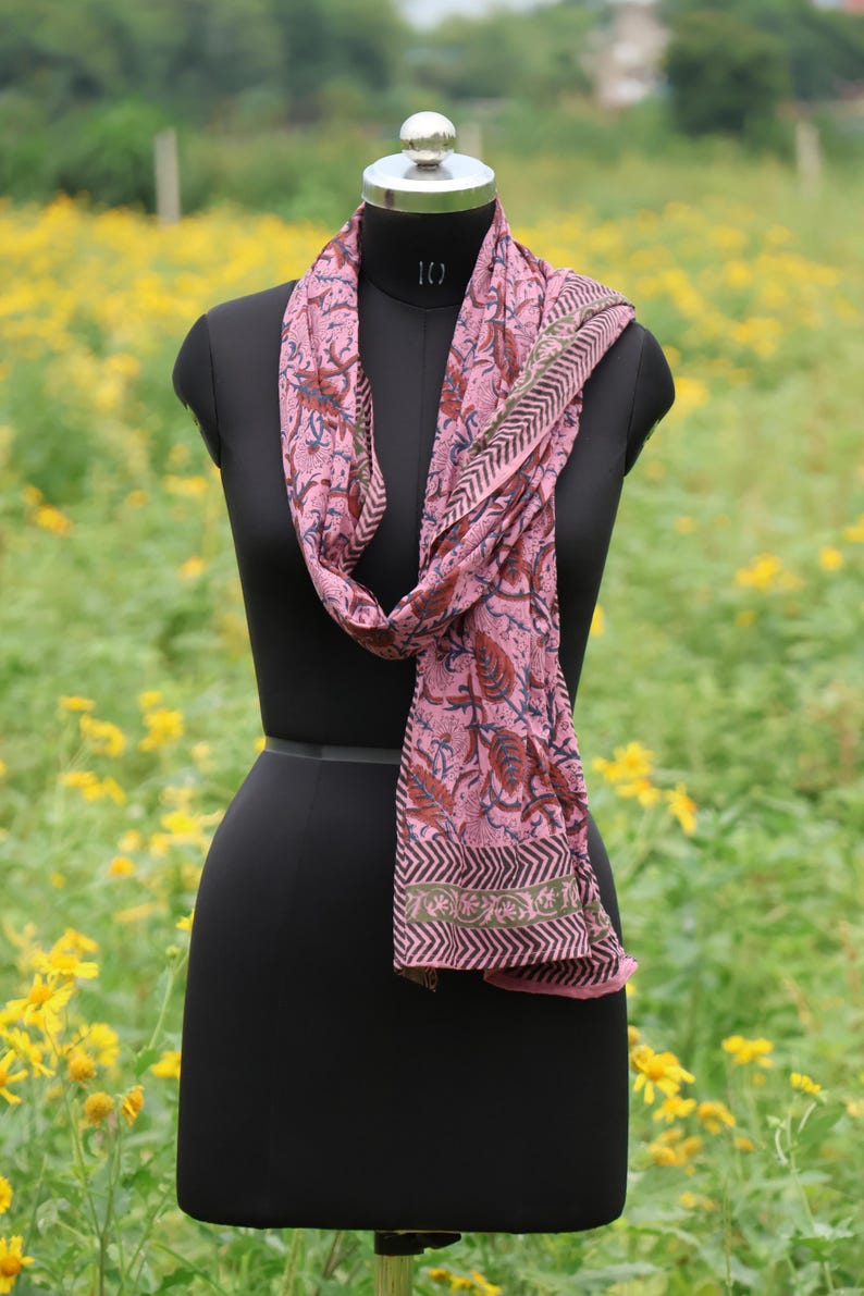 May include: A pink scarf with a floral pattern and a geometric border is draped on a black mannequin. The scarf features a repeating design of leaves and flowers in shades of red, blue, and brown. The background is a field of yellow flowers.