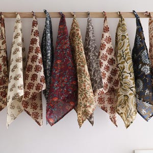 Puede incluir: Una colección de diez toallas de tela estampadas en varios colores, incluyendo crema, marrón, burdeos y azul marino. Cada toalla presenta un diseño floral y cuelga de una barra de madera. Las toallas están hechas de tela suave.