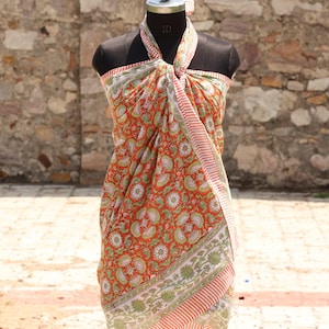 May include: A vibrant orange sarong with a floral pattern in green and white, draped on a mannequin. The sarong has a striped border in red and white. The fabric is lightweight and appears to be cotton.