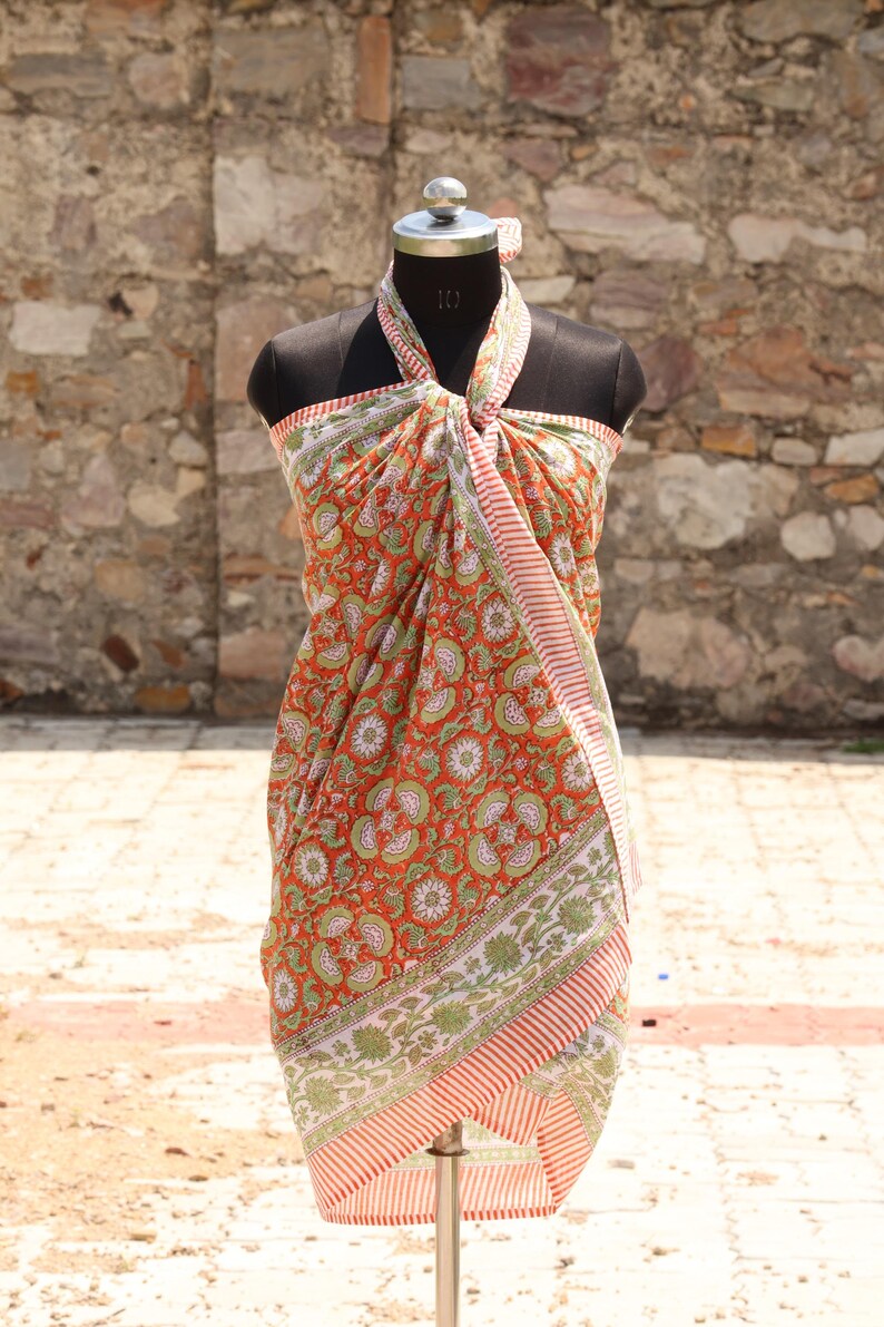 May include: A vibrant orange sarong with a floral pattern in green and white, draped on a mannequin. The sarong has a striped border in red and white. The fabric is lightweight and appears to be cotton.