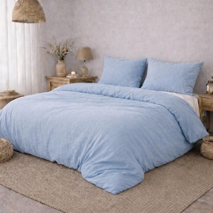 May include: A pale blue bed set, including a duvet cover and two pillowcases, on a bed with a beige rug. The room has a neutral colour palette with wooden beams and a woven pendant light.