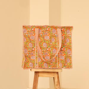 May include: An orange tote bag with a floral pattern of white and green flowers and leaves. The bag has two handles and is sitting on a small wooden stool. The bag's edges are trimmed with a white and orange stitch.