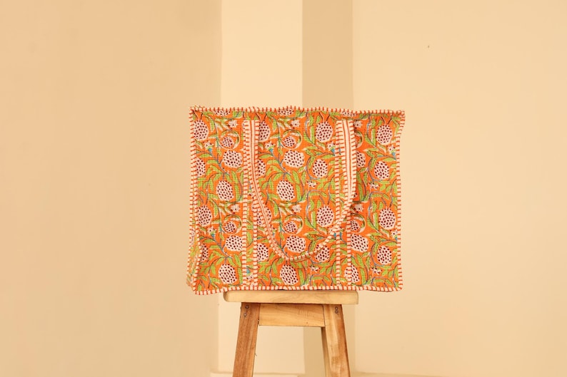 May include: An orange tote bag with a floral pattern of white and green flowers and leaves. The bag has two handles and is sitting on a small wooden stool. The bag's edges are trimmed with a white and orange stitch.