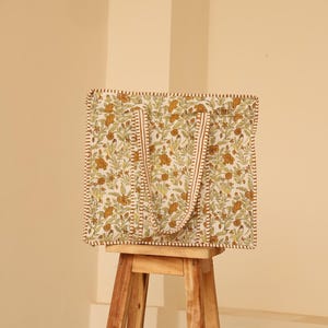 May include: A square tote bag with a floral pattern in shades of green and brown on a cream background. The bag has matching handles and trim. The bag is sitting on a wooden stool.