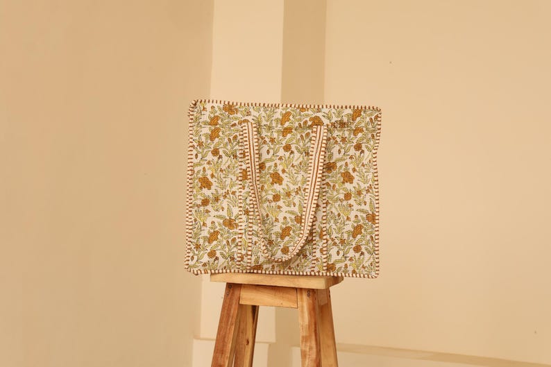 May include: A square tote bag with a floral pattern in shades of green and brown on a cream background. The bag has matching handles and trim. The bag is sitting on a wooden stool.